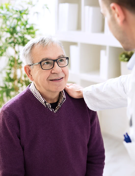 an elderly man in a purple sweater smiling and looking up at a doctor who places his hand on the mans shoulder.